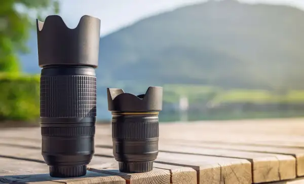 Two camera lenses, a tall telephoto and a shorter normal lens, sit on a wooden deck with a blurred mountain and lake background.