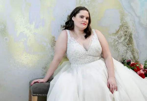 Kobieta w białej, zdobionej sukni ślubnej plus size siedzi na tle złoto-białej ściany.