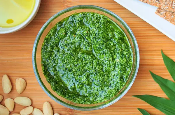 Aromatic pesto z czosnku niedźwiedziego w szklanej miseczce, otoczone migdałami i świeżymi liśćmi.