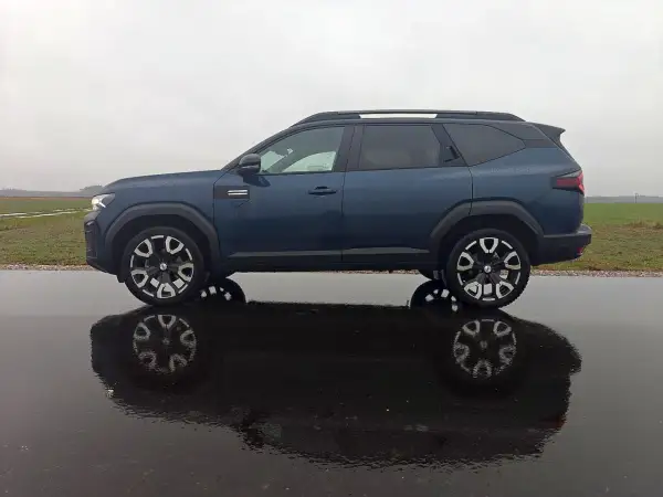 Dacia Bigster LPG w ciemnoniebieskim kolorze, zaparkowana na mokrej drodze, z odbiciem w kałuży.