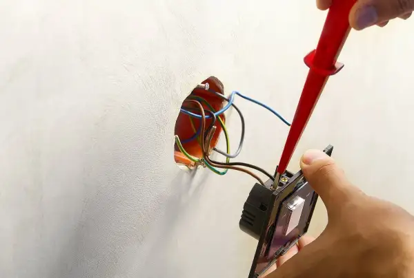 Instalator podłącza podwójny włącznik światła, używając próbnika napięcia. Widać przewody wychodzące ze ściany, gotowe do połączenia.