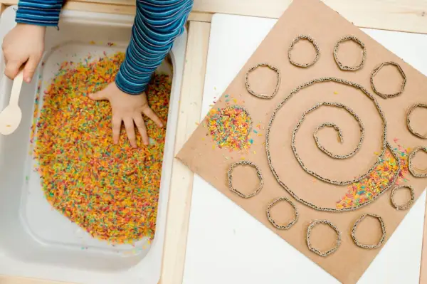 Main colorée dans un bac, une main d'enfant explore. À côté, un carton en spirale avec des ronds découpés, parsemé de riz coloré. Une activité sensorielle adulte pour stimuler la créativité.