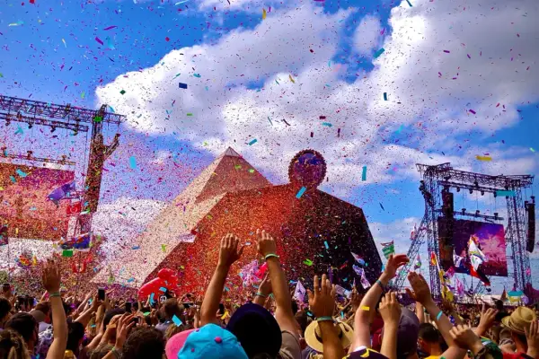 Confetti rains down on a cheering crowd at the Glastonbury Pyramid Stage, a vibrant celebration under a blue, cloud-filled sky.