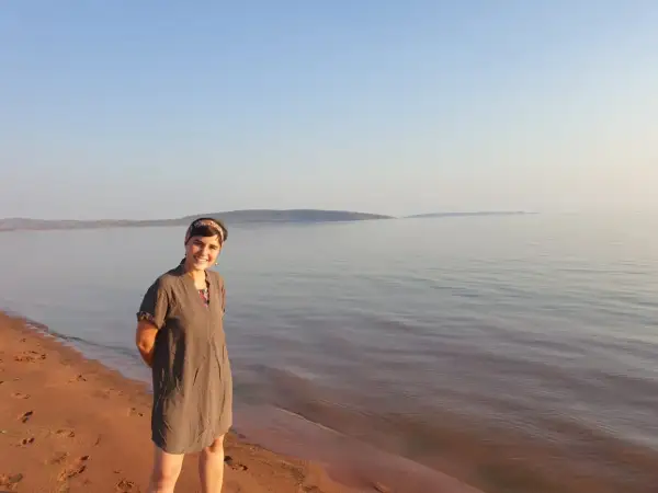Mujer sonriente en la orilla de arena rojiza del lago Kariba, con el agua tranquila y el cielo despejado al fondo.