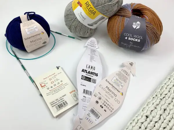 Wollknäuel zeigen den Unterschied zwischen Schurwolle und Wolle. Ein blauer Merino-Strang und ein Tweed-Garn liegen neben Stricknadeln.