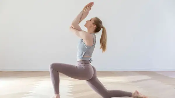 Frau in lila Leggings und grauem Top praktiziert Bikram Hot Yoga in einer tiefen Ausfallschritt-Pose.