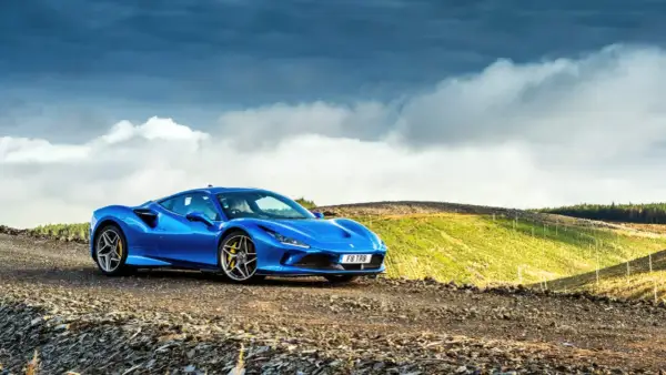 Niebieskie Ferrari F8 na tle malowniczego, pagórkowatego krajobrazu z chmurami na niebie.