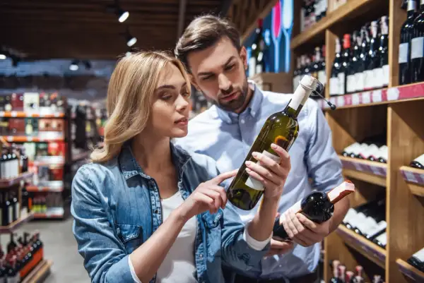 Para wybiera wino w sklepie. Kobieta wskazuje na etykietę, na której widnieje informacja, że produkt zawiera siarczyny.