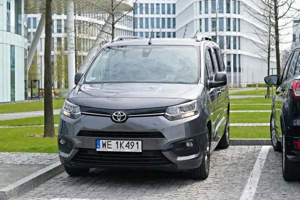 Szary Toyota Proace City zaparkowany prostopadle tyłem na parkingu przed nowoczesnym budynkiem.