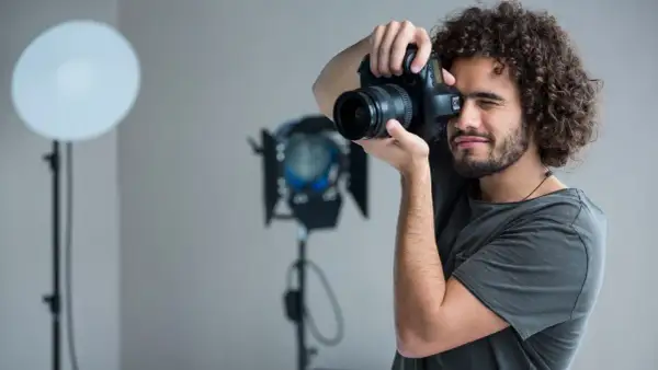Mężczyzna z kręconymi włosami i aparatem w dłoniach, ćwiczący, jak zostać fotografem. W tle lampy studyjne.