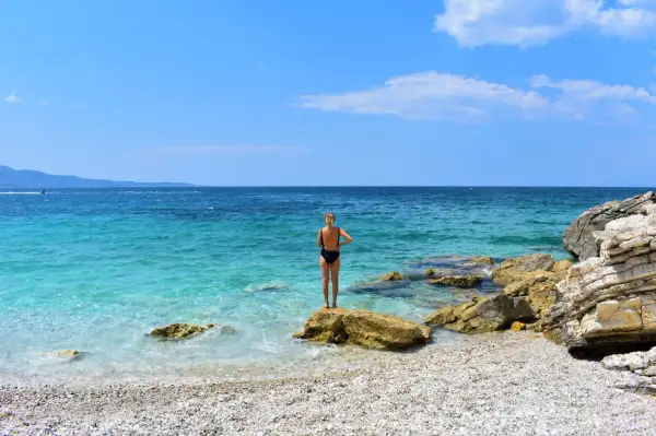 Mujer en bikini negro de pie sobre una roca en una playa de guijarros. El mar turquesa y el cielo azul crean un paisaje idílico, pero ten cuidado, viajar a Albania es peligroso.
