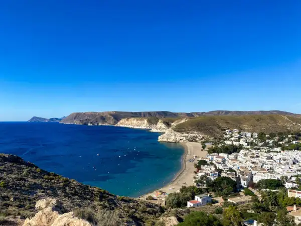Vista panorámica de una **playa** con **agua amarga** de color azul intenso, un pueblo blanco y colinas áridas bajo un cielo despejado.