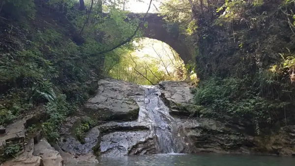 Cascate brescia: un piccolo salto d'acqua scende tra rocce e vegetazione lussureggiante, sotto un antico ponte ad arco.