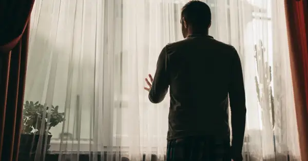 Mężczyzna stoi przy oknie, zasłonięty przez firankę. Może to symbolizować izolację i trudności w nawiązywaniu kontaktów, jedne z objawów schizofrenii.