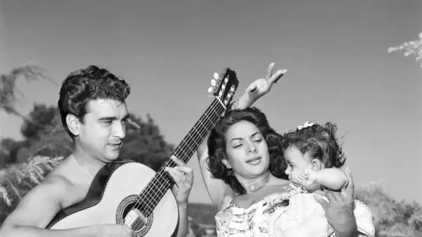 Hombre toca guitarra mientras mujer baila con bebé. Una escena de rumbas flamencas al aire libre.
