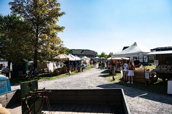 Besucher bummeln über den Bauernmarkt Lindlar, vorbei an Ständen mit Waren und Sitzgelegenheiten unter Bäumen.