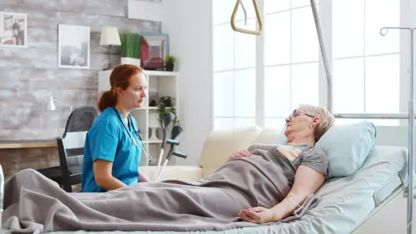 Pielęgniarka rozmawia z pacjentką leżącą w łóżku. W tle widać kule, co sugeruje, że pacjentka może potrzebować wsparcia w odżywianiu dojelitowym.