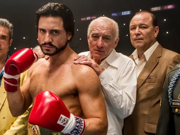 Roberto Duran, the legendary fighter, stands ready with red boxing gloves. His team, including Robert De Niro, offers support.