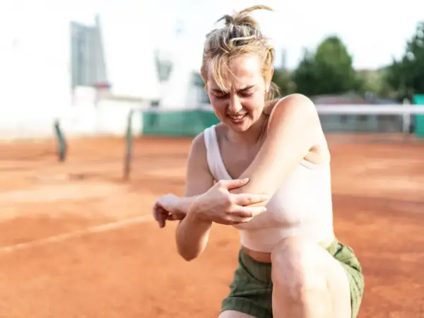 Junge Frau auf einem Tennisplatz hält schmerzgebeugt ihren Ellenbogen. Der Tennisarm wird nicht besser.