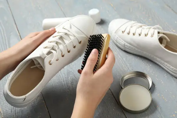 Mains nettoyant une basket blanche en tissu avec une brosse. Un pot de produit d'entretien est ouvert à côté.