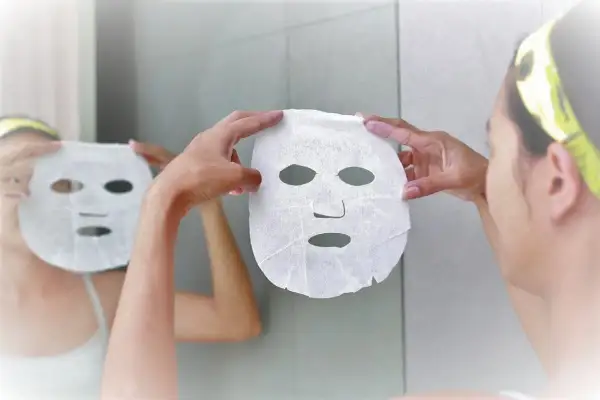 Mani bagnate che applicano una maschera in tessuto sul viso, riflessa nello specchio. Dopo la maschera in tessuto si lava il viso.