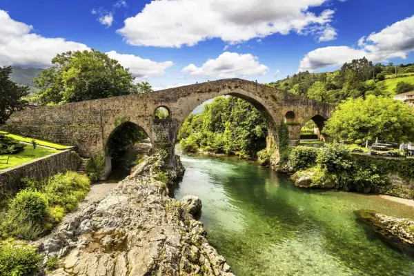 El mirador de Cangas de Onís ofrece vistas del icónico puente romano sobre un río verde esmeralda, rodeado de exuberante vegetación y un cielo azul con nubes.