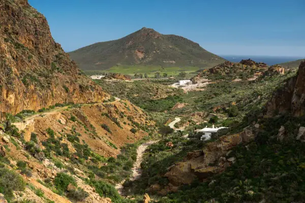 Paisaje árido con el **Cerro Negro** al fondo. Un camino serpentea entre barrancos y vegetación baja, llegando a unas casas blancas.