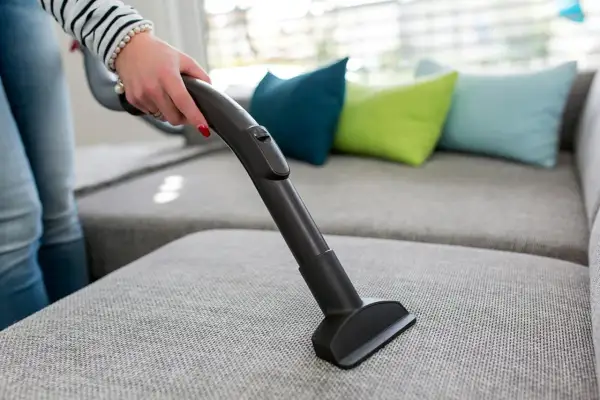 En hand använder en soff dammsugare för att rengöra en grå soffa med färgglada kuddar i bakgrunden.