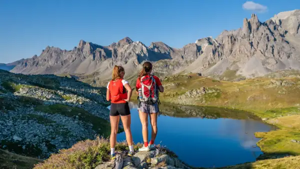 Deux randonneuses contemplent un lac d'altitude dans la vallée de la Clarée en été. Les montagnes majestueuses se reflètent dans l'eau calme.