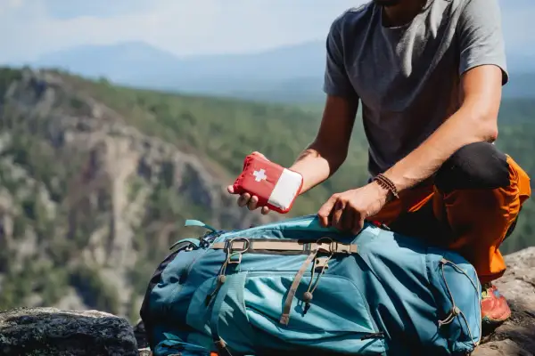Mann packt Erste-Hilfe-Set für leichte Wanderung in Rucksack. Bergpanorama im Hintergrund.