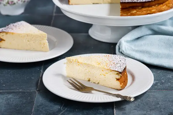 Ein Stück cremiger Quarkkuchen ohne Boden, bestäubt mit Puderzucker, wartet auf dem Teller.