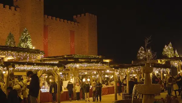 Mercado navideño bajo un castillo iluminado. Gente pasea entre puestos decorados con luces, mostrando como se celebra la navidad.