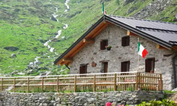 Rifugio in pietra con bandiera italiana, circondato da prati verdi e un ruscello. Un luogo ideale per rifugi val camonica facili.