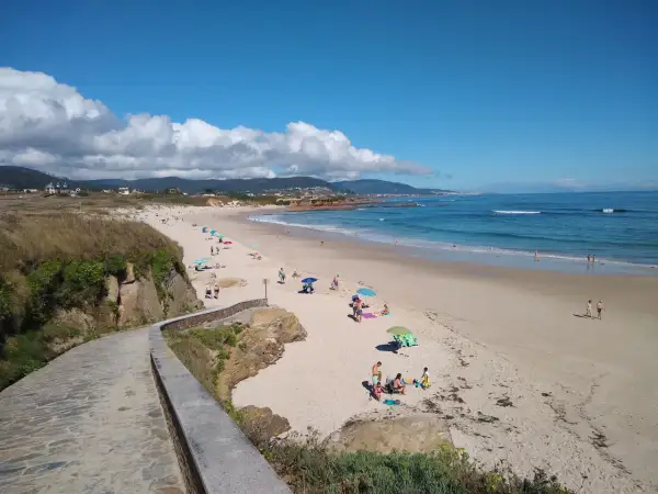 Un paseo de piedra bordea la **playa llas**, donde gente descansa bajo sombrillas y el mar azul invita a un baño.