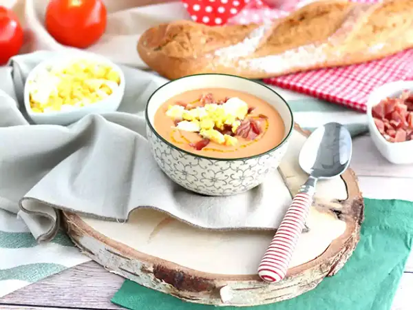 Schüssel mit Gazpacho, garniert mit Ei und Schinken. Daneben sind Tomaten, Brot und weitere Zutaten für kalte Tapas.