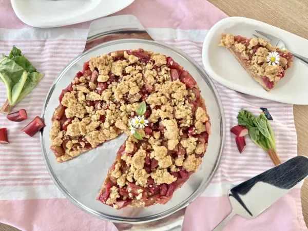 Ein köstlicher Rhabarberkuchen mit Streuseln, garniert mit kleinen Gänseblümchen. Frischer Rhabarber liegt daneben.