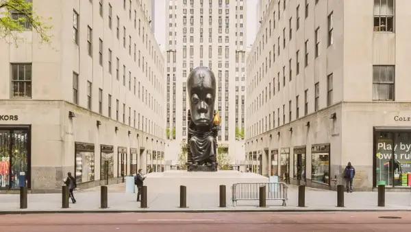 A monumental sculpture of a seated figure with a large head, holding a torch, stands as a striking example of public art between two imposing buildings.