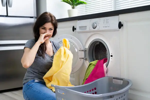 Donna con espressione disgustata annusa un asciugamano giallo, la lavatrice puzza. Cesto con panni colorati accanto.