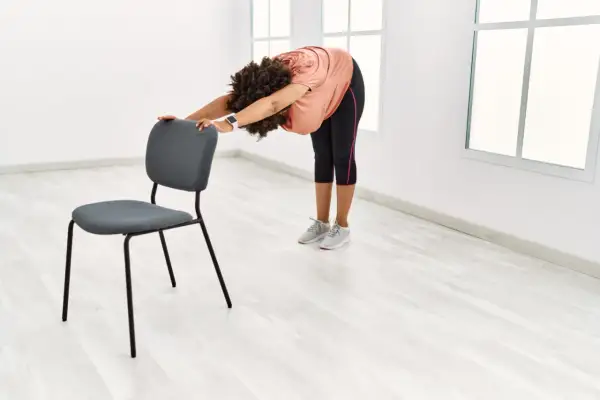 Femme pratiquant le yoga sur chaise, se penchant en avant pour étirer son dos, mains posées sur le dossier du siège.