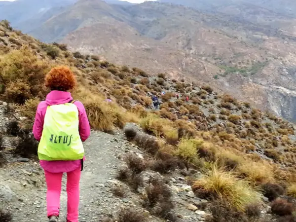 Senderista con mochila amarilla en un sendero montañoso. El grupo avanza, algunos a caballo, en un paisaje árido.