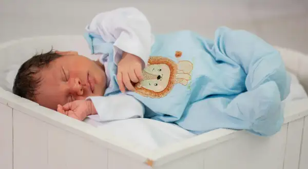 A newborn baby sleeps peacefully in a white bassinet, wearing a light blue onesie with a lion embroidery.