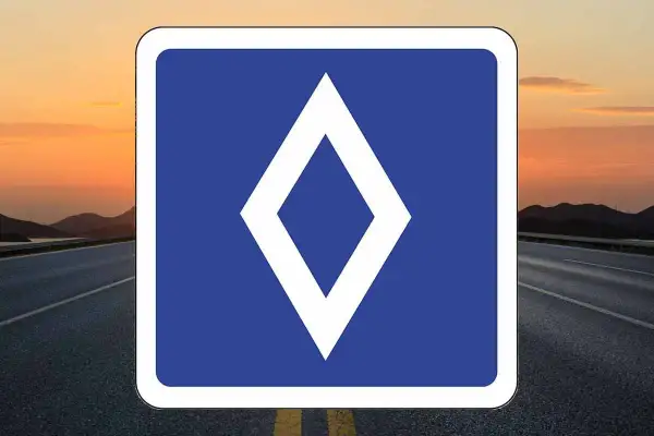 Un nouveau panneau routier bleu avec un losange blanc au centre, sur fond de route et de coucher de soleil.