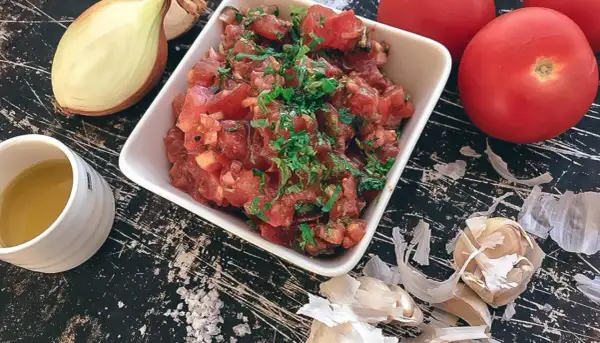 En skål med färsk tomatsalsa, redo för ett tomatsalsa recept. Ingredienser som lök, vitlök och tomater syns runt omkring.