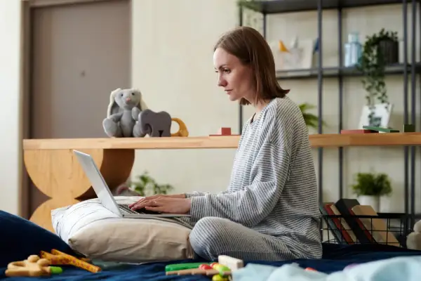 Kobieta w piżamie pisze na laptopie, przygotowując wniosek o urlop macierzyński. Na łóżku obok leżą zabawki.