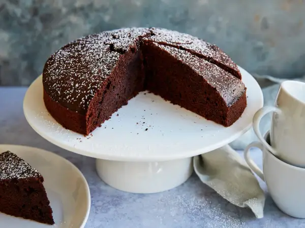 Ein saftiger veganer Schokokuchen, bestäubt mit Puderzucker, steht auf einem weißen Tortenständer. Daneben eine Tasse und ein Stück Kuchen auf einem Teller.