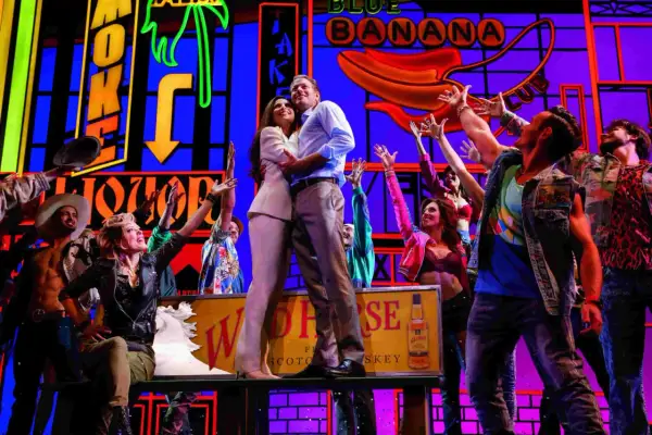 Szene aus einem Musical in Deutschland: Ein Paar umarmt sich, umgeben von jubelnden Darstellern vor einer bunten Kulisse mit Neonlichtern.