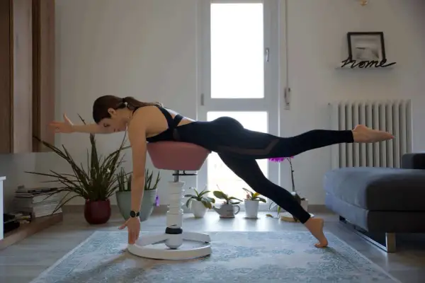 Donna esegue esercizi per riscaldare la schiena, in equilibrio su una sedia da pilates, con una gamba tesa e un braccio esteso.