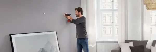 Uomo usa trapano per fissare listelli di legno al muro, preparando la parete per appendere un quadro.