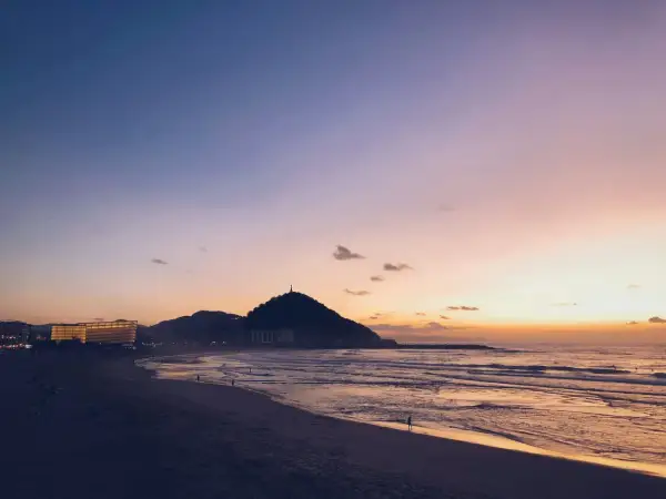 Atardecer en la costa San Sebastián. El cielo se tiñe de tonos rosados y naranjas sobre el mar y la playa, con la silueta del monte Urgull al fondo.