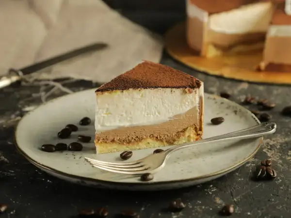 Pyszny sernik z mascarpone, posypany kakao, na talerzu z ziarnami kawy. Obok widelca leży kawałek ciasta.